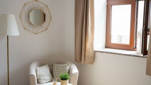Een kamer met een beige bank, een ronde tafel met een decoratieve schaal en een plant, een geometrisch spiegel aan de muur en een raam met gordijnen.
