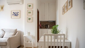 Een woonkamer met een beige bank, een witte bijzettafel met een plant, en een witte stoel. Er is een keukenhoek met witte kasten en een betegelde achterwand.