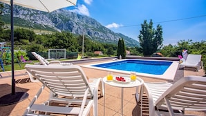 A pool area with lounge chairs and a table with drinks, surrounded by mountains and greenery.