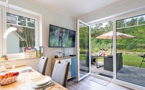 A modern dining area with a television, a dining table set with plates and glasses, and a view of the outdoors through large glass doors.