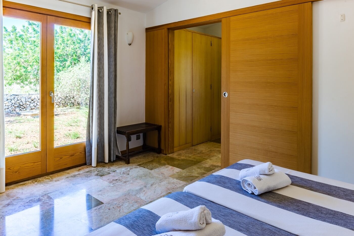 A room with two beds, a wooden wardrobe, a bench, and a sliding glass door leading to an outdoor area.