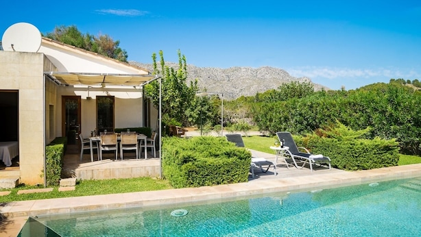 Un espace aménagé au bord de la piscine, avec un coin repas et des chaises longues, entouré de verdure et dominé par un paysage montagneux.