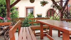 An outdoor patio with wooden benches and a table, surrounded by greenery and a rustic building.