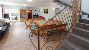 A spacious living area with a wooden dining table, comfortable seating, a staircase with a decorative railing, and a kitchen in the background.
