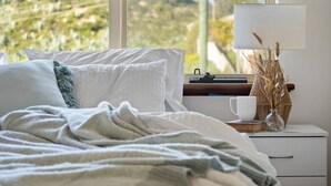 A bedroom with a bed, a bedside table with a lamp, a cup, and a vase with dried plants. - Absolute Beachfront Escape · Ocean Views · Family Luxury (Halls Head)
