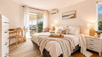 A bedroom with a bed, bedside tables, a desk, a chair, a balcony with curtains, and a painting on the wall.