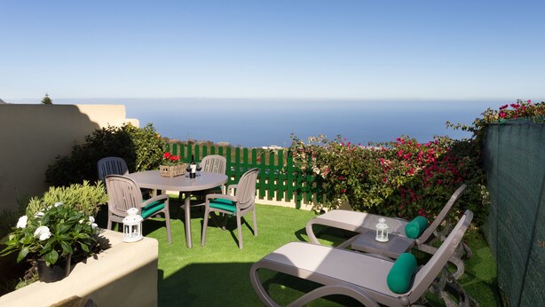Un patio con una mesa y sillas, vista al mar y un seto verde.