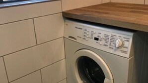 Un lavadero pequeño y moderno, con lavadora y secadora integradas, paredes con azulejos y una ventana que proporciona luz natural.
