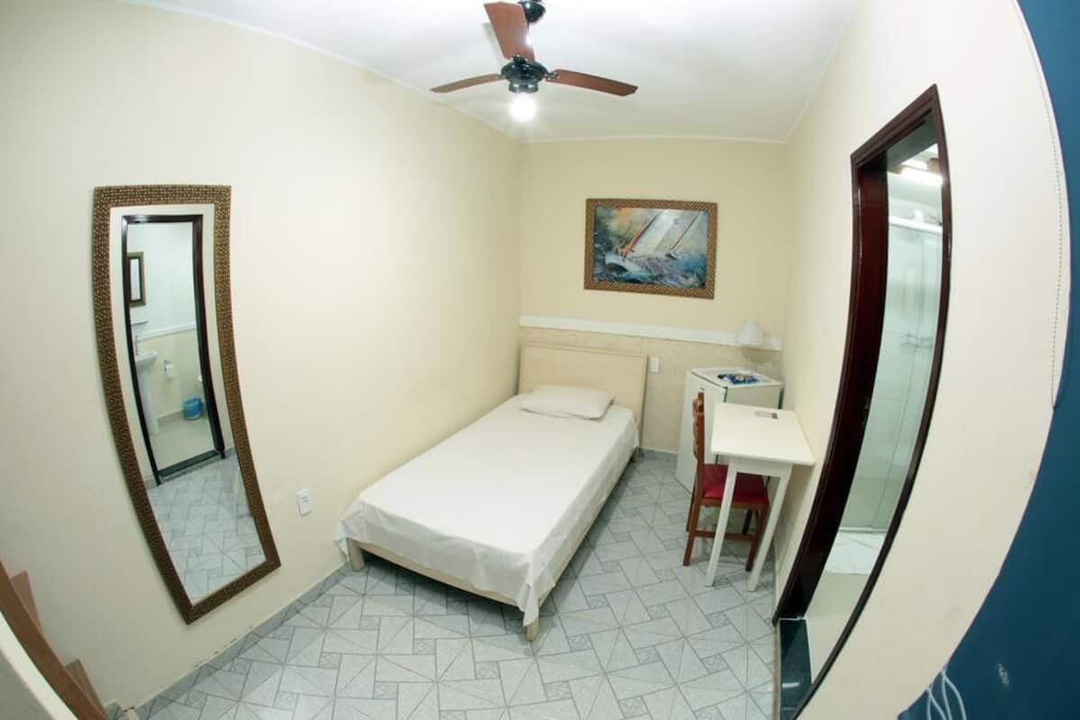 Una habitación pequeña con cama individual, ventilador de techo, mesita de noche, una silla y un espejo.