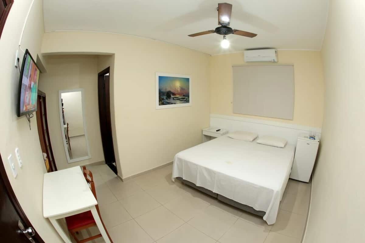 Un dormitorio con cama, escritorio, silla, ventilador de techo, televisor montado en la pared y un cuadro.