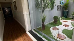 A hallway with wooden flooring, a glass partition, and a small garden area with artificial grass and rocks. - Hotel Colonial (Porto Ferreira)