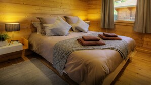 Un dormitorio de cabaña de madera acogedor, con cama, almohadas, una mesita de noche, una lámpara y una ventana con cortinas.
