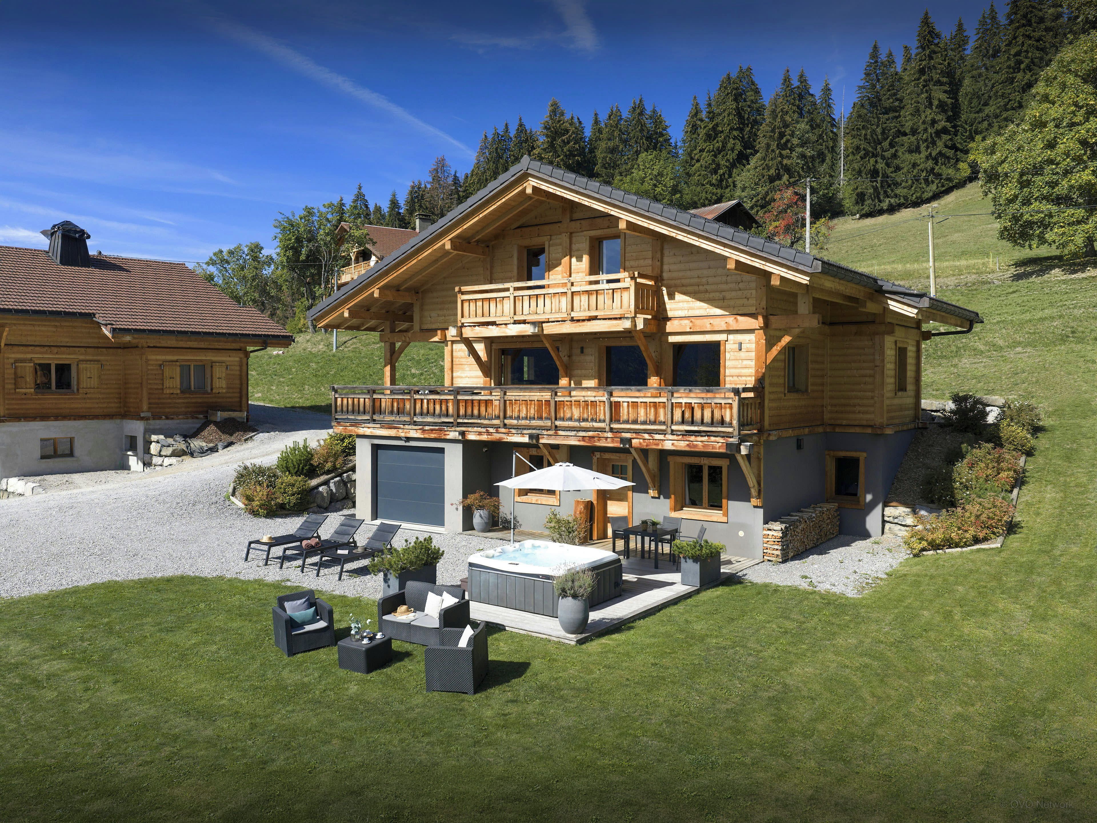 A two-story wooden chalet with a balcony, outdoor seating, and a hot tub.
