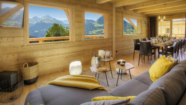 Interior de una cabaña de madera con comedor, sofá y amplios ventanales con vistas a las montañas.