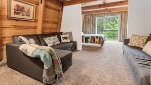 Interior acogedor de una cabaña con detalles de madera, un sofá, una cama con edredón de motivos forestales y un ventanal que ofrece vistas al exterior.