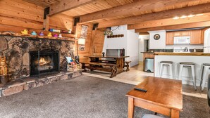 Una sala de estar acogedora con chimenea de piedra, techo de madera y una zona de cocina con taburetes de bar.