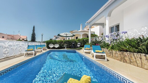 Ein Swimmingpool mit Liegestühlen und Sonnenschirmen, umgeben von einem weißen Gebäude mit einer gefliesten Terrasse.