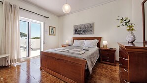 Ein Schlafzimmer mit einem hölzernen Bett, einem Nachttisch mit Lampe, einer Kommode und Blick ins Freie durch eine Schiebetür.