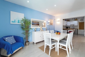 Una sala moderna con un sofá azul, una mesa de comedor con sillas blancas, una zona de cocina y una planta grande en maceta.