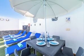 Un espacio al aire libre iluminado por el sol, con sillas de descanso, una mesa lista para comer y un gran paraguas que ofrece sombra.