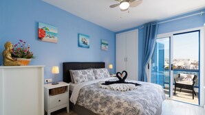Un dormitorio con una cama, mesitas de noche, una lámpara de pared, una estatua decorativa de Buda y una ventana con vistas a edificios.