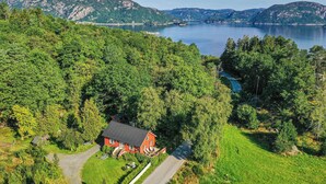 Ein rotes Haus mit befestigter Auffahrt und kleinem Rasen, umgeben von dichtem Grünwald und einem See im Hintergrund.