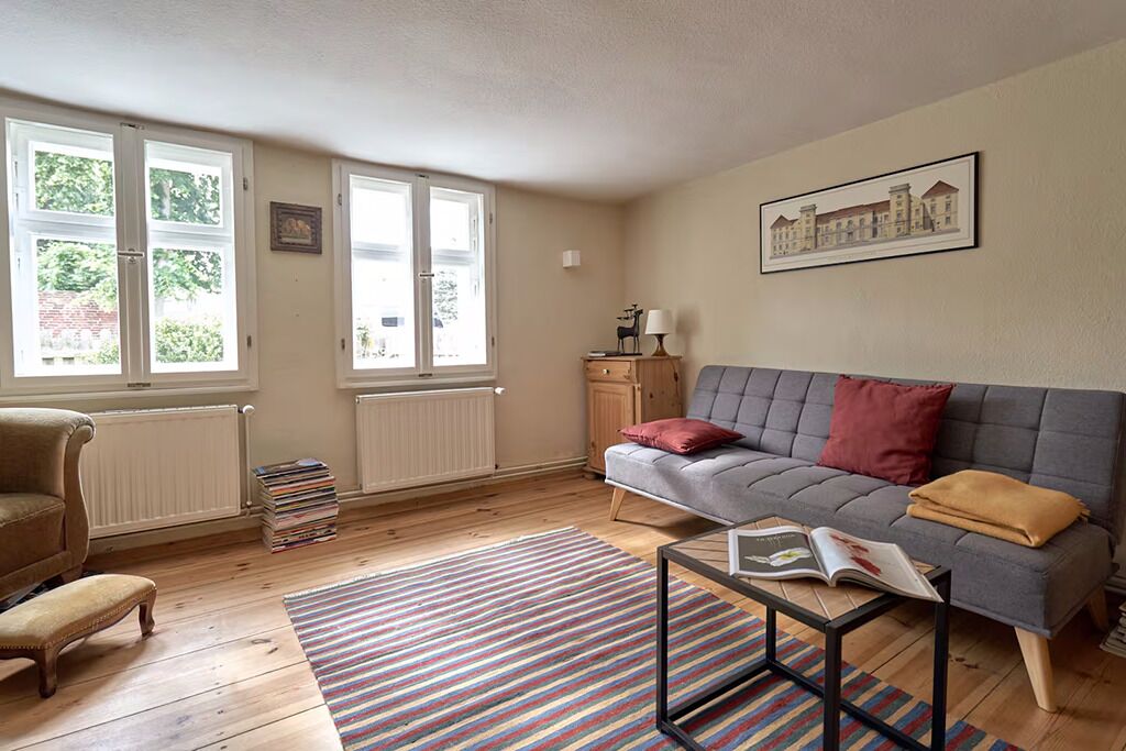 Ein Wohnzimmer mit einer Couch, einem Sessel und einem Couchtisch mit einem Buch. Es gibt Fenster, eine Heizung und ein gerahmtes Bild an der Wand.