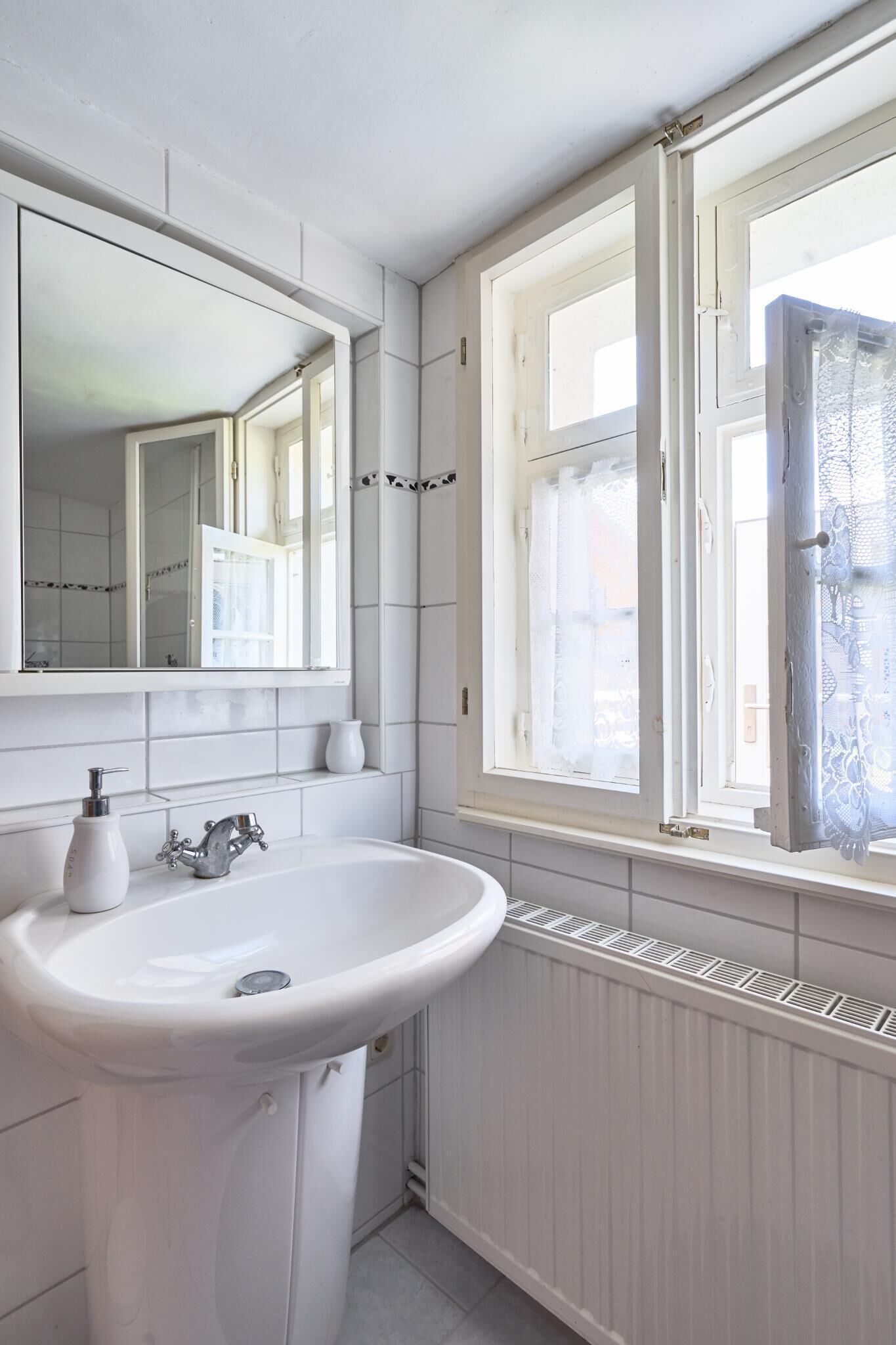A bathroom with a white sink, mirror, and window with lace curtains.