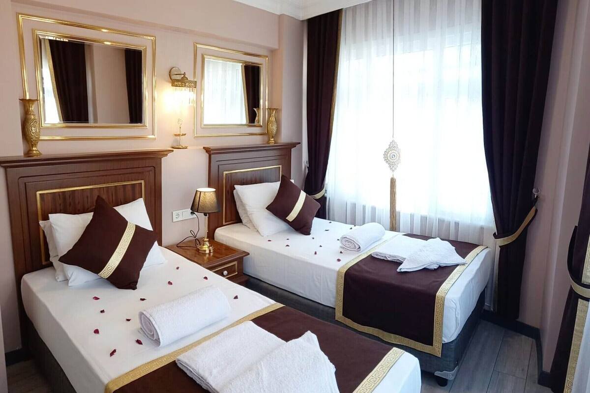 A hotel room with two beds, a large window with curtains, a mirror, and a bedside table with a lamp.