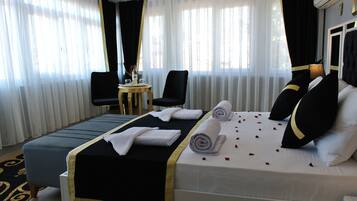 A hotel room with a large bed, a small round table with a chair, and a window with sheer curtains.