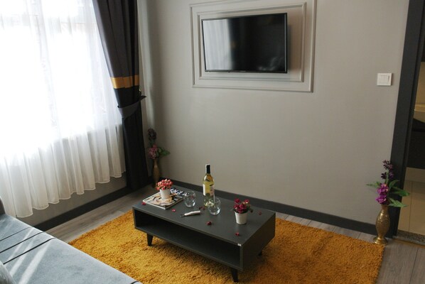 A modern living room with a dark coffee table, a flat-screen TV mounted on the wall, a sofa, and a yellow rug. - Emirhan Guest House & Suites (Istanbul)
