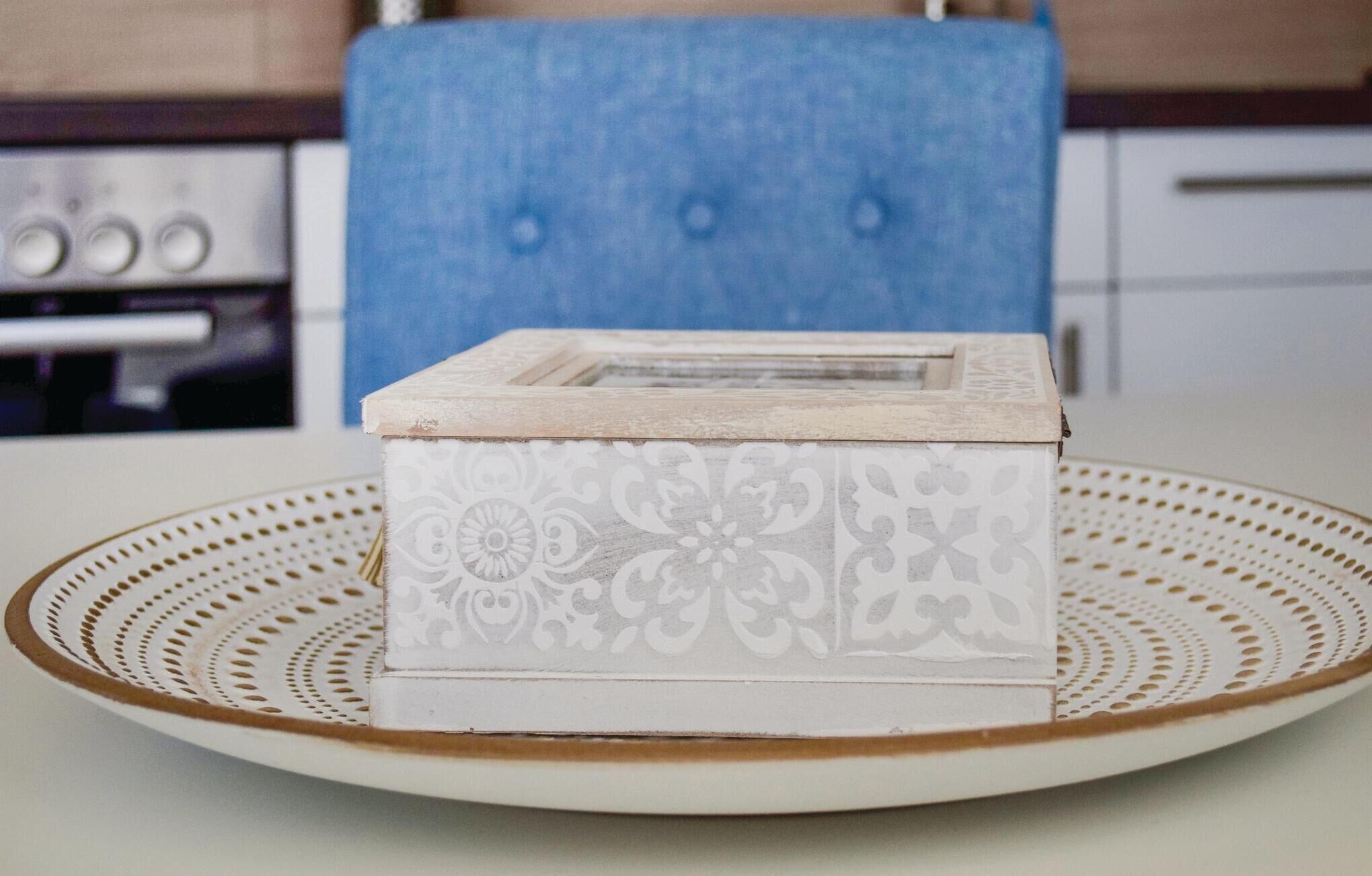 A white decorative box with a floral pattern on a round table with a brown edge, in a kitchen setting with a blue chair and a stove in the background.
