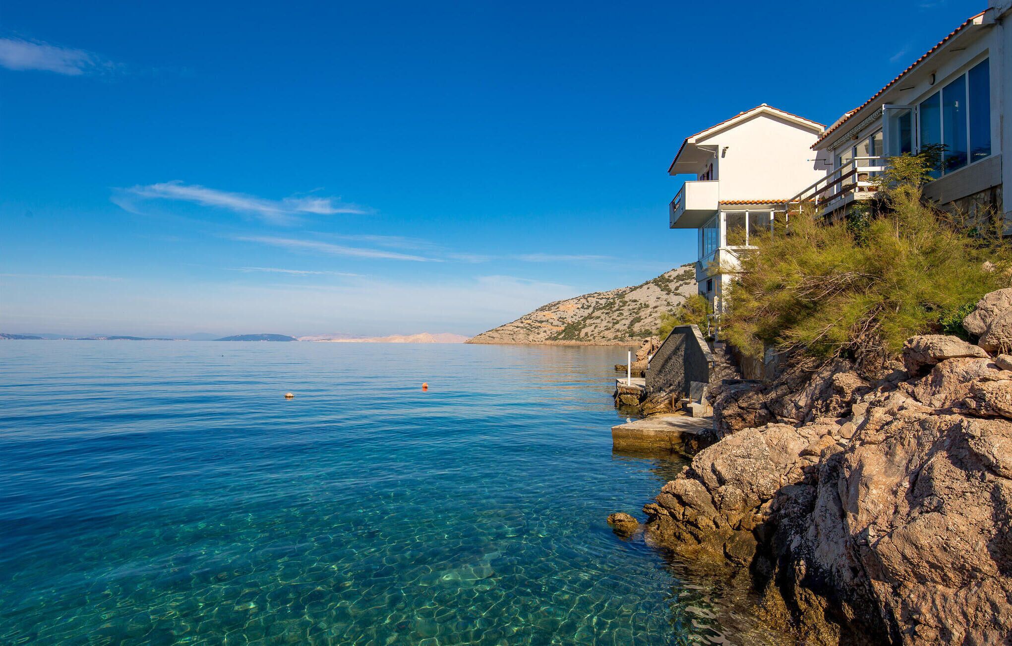 Um imóvel costeiro com águas azuis cristalinas, terreno rochoso e um edifício branco com varandas com vista para o mar.