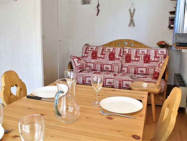 A dining area with a wooden table set for two, a patterned sofa, and a small kitchen area in the background.