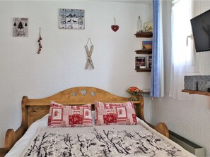 A bedroom with a wooden bed, patterned bedding, a wall with a picture and a heart decoration, and a shelf with books and a bottle. - Studio for 4 near shops and slopes in Risoul (Risoul)