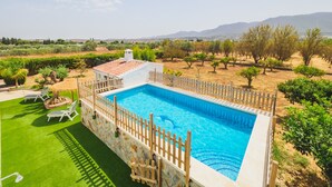 Una piscina rettangolare con recinzione bianca e muro in pietra, circondata da un prato curato e affacciata su un vigneto e montagne in lontananza.
