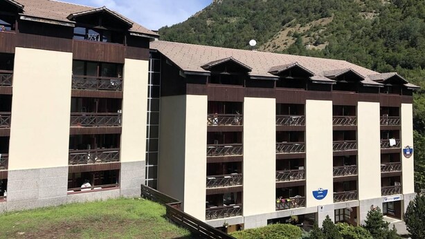 Un hotel de varios pisos con balcones, una señalización de pista de esquí y un paisaje montañoso de fondo.