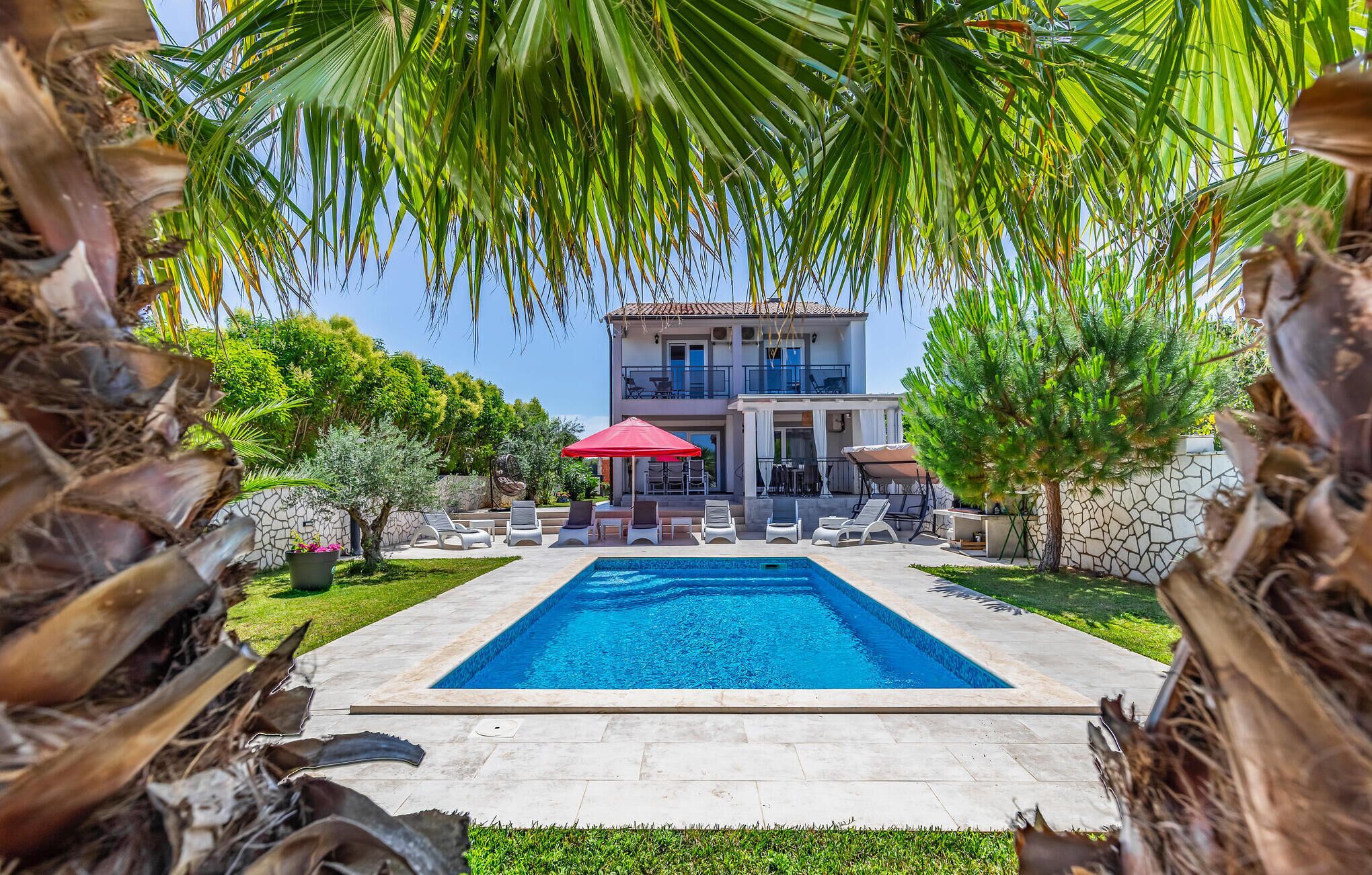 A swimming pool with lounge chairs and umbrellas, surrounded by palm trees and a two-story building with balconies.