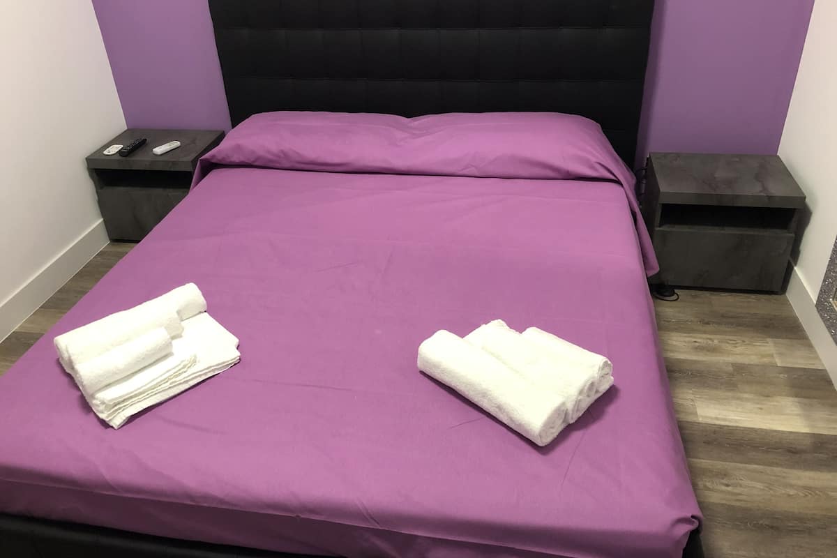 A bed with a purple bedspread and two white towels folded on it. There are two nightstands, each with a remote control and a phone.