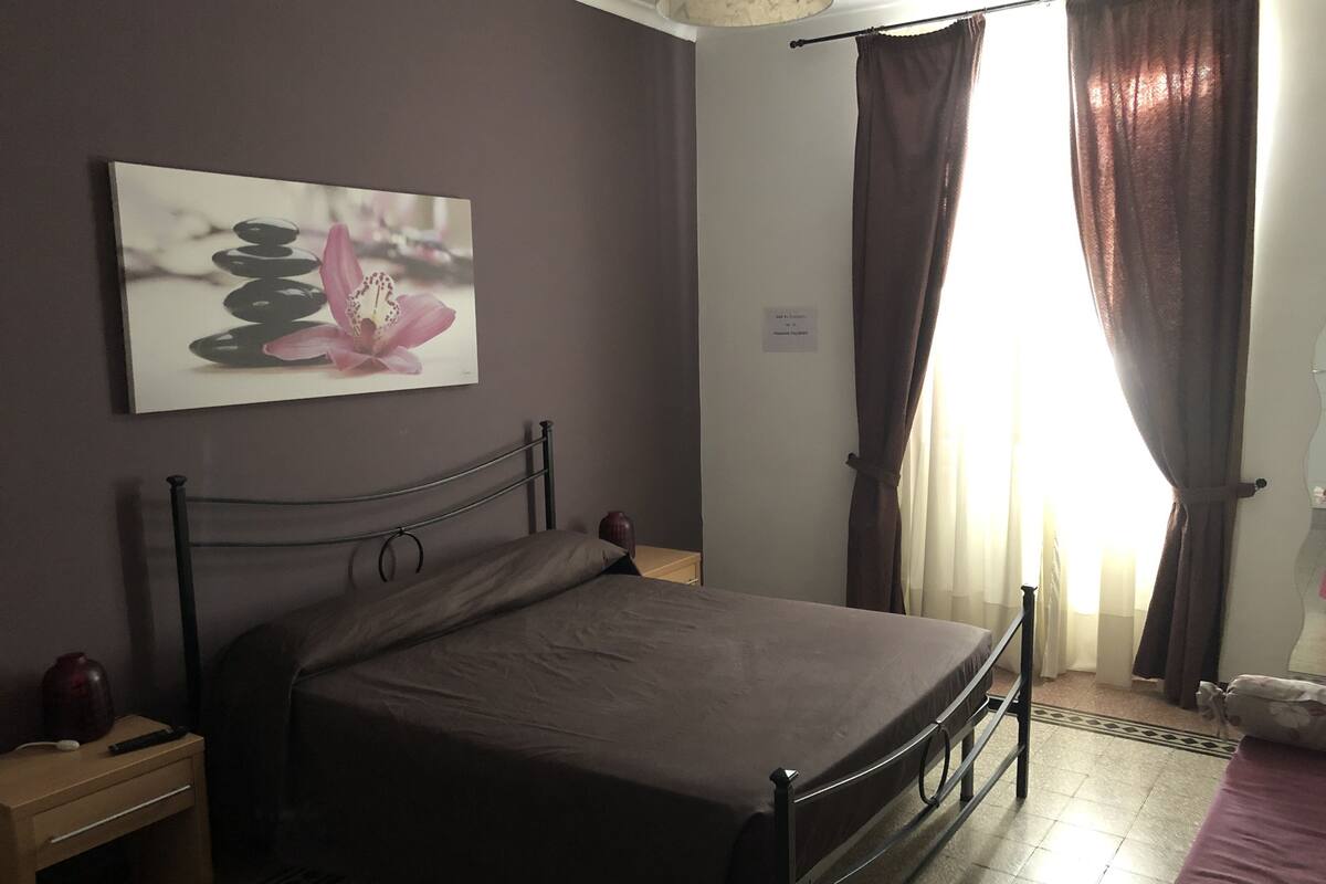 A bedroom with a bed, a nightstand, a wall-mounted painting, and a window with curtains.