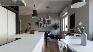 Moderni keittiö, jossa on keskeinen saareke, riippuvalaisimet sekä ruokailutila, jossa on valkoinen pöytä ja tuolit.