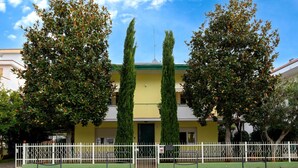 A building with a yellow facade, three tall cypress trees in front, and a white fence with a bench in the foreground. - Nice apartment in Misano Adriatico (RN) (Misano Adriatico (RN))