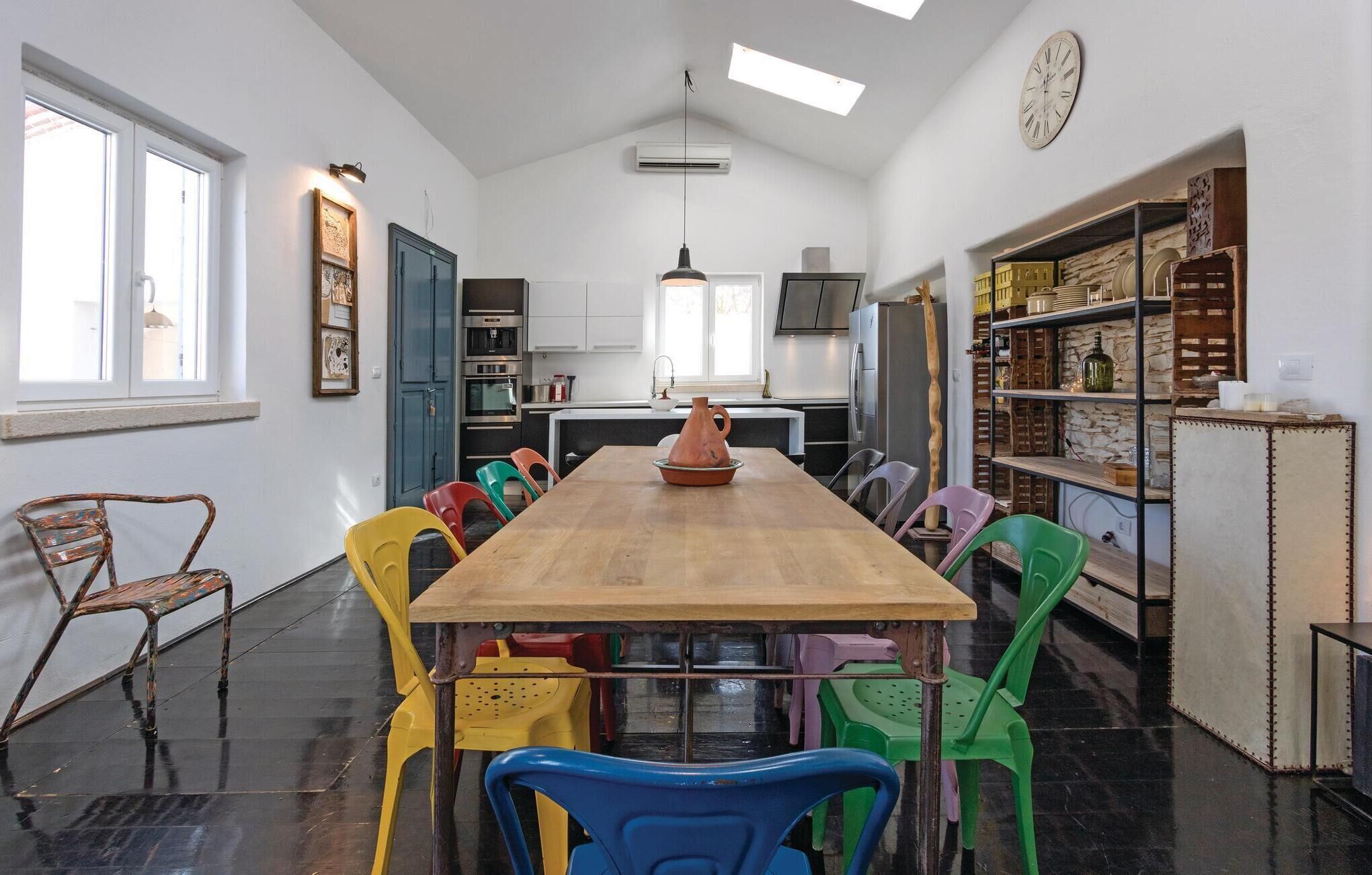 Un coin repas avec une table en bois et des chaises en plastique de couleurs vives, une cuisine équipée d’appareils électroménagers modernes et une bibliothèque contenant divers objets.