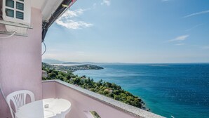 Un balcon avec une table et des chaises blanches, offrant une vue sur la côte, avec une mer bleue et limpide et une terre lointaine à l’horizon.