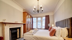 A bedroom with a large bed, a fireplace, a chandelier, and a view of buildings through the window. - Victorian townhouse near International Conference Centre / Kings Quarter (Harrogate)