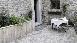 Un edificio de piedra con una cerca de madera, una puerta y una ventana adornada con flores. Hay una mesa con mantel blanco y sillas en el exterior.