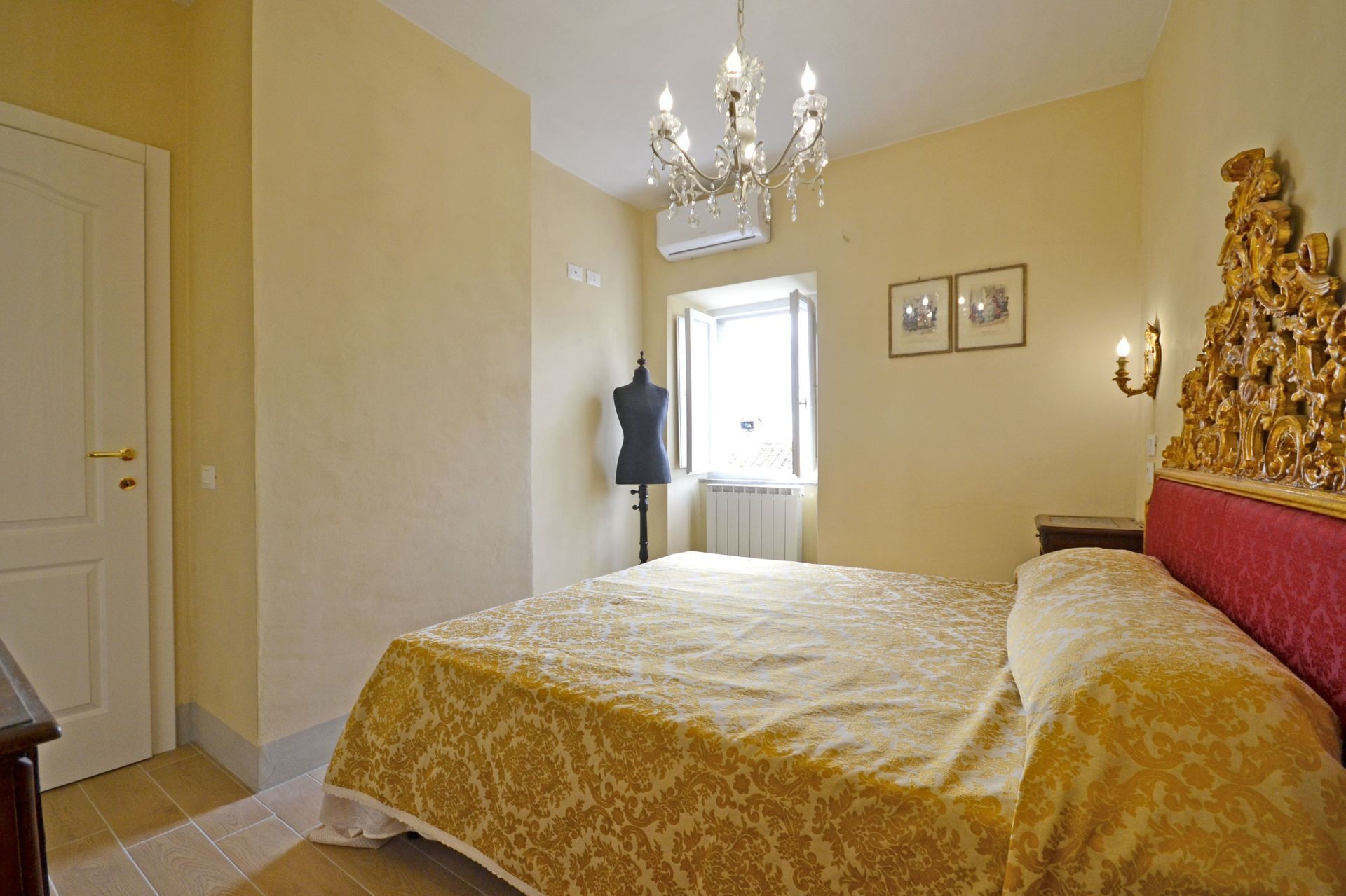 A bedroom with a bed, a chandelier, a dresser, and a window with an open door.