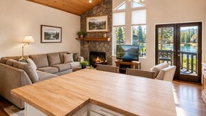 A spacious living area with a stone fireplace, a large wooden dining table, and a view of a lake through glass doors. - Lakefront Viking Oasis Condo - Lakefront, Indoor Pool, Hot Tub (Big Bear Lake)