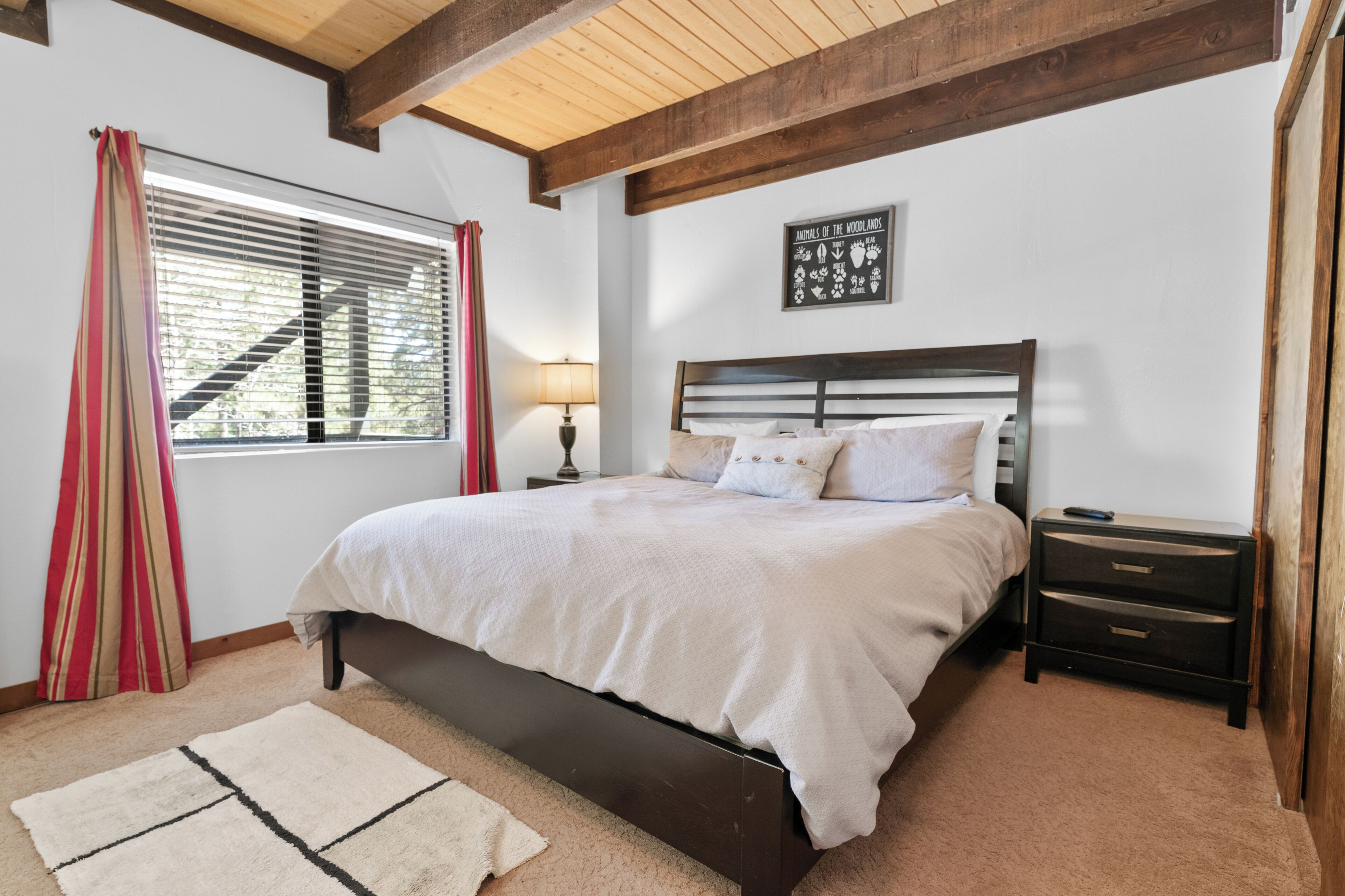 A bedroom with a bed, a nightstand, a lamp, a window with curtains, and a wall art piece.