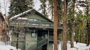 Een chalet met een balkon en een trap naar de ingang, omgeven door hoge bomen en bedekt met sneeuw.